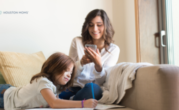 Mom sitting on couch looking at her phone while child draws next to her