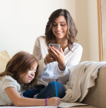 Momeaze: An App that Manages the Mental Load of Motherhood Mom sitting on couch looking at her phone while child draws next to her