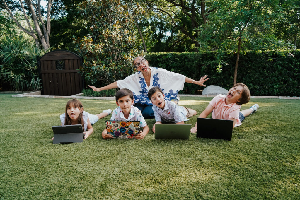 A woman sits in the grass making a silly face with four kids and their laptops